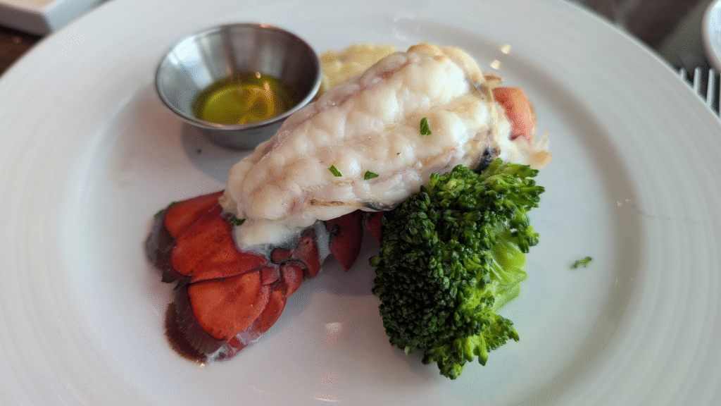 A perfectly cooked, butterflied lobster tail served with a side of bright green broccoli and mashed potatoes, with drawn butter, photographed on a white plate during the ship's formal night.
