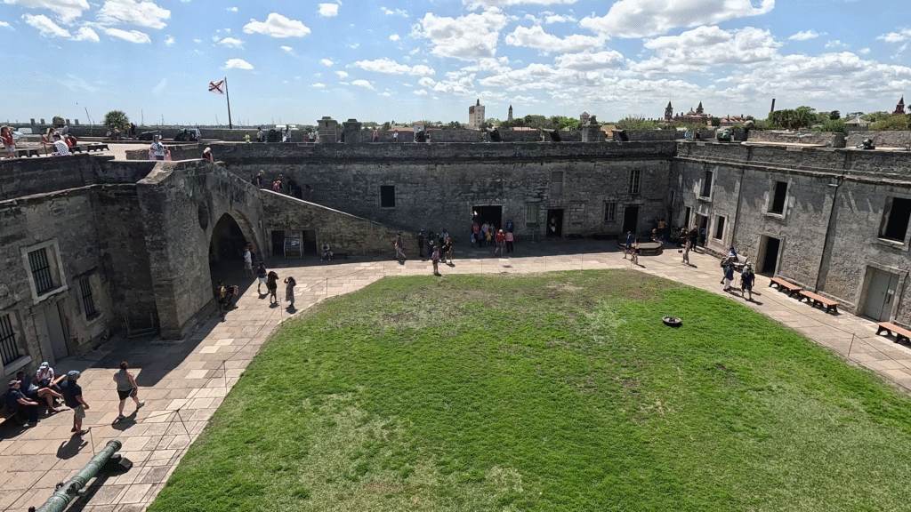 High-angle view of the Castillo de San Marcos interior courtyard and Plaza de Armas in St. Augustine, Florida, featuring the coquina stone walls, a Spanish flag flying, and the green lawn with visitors.