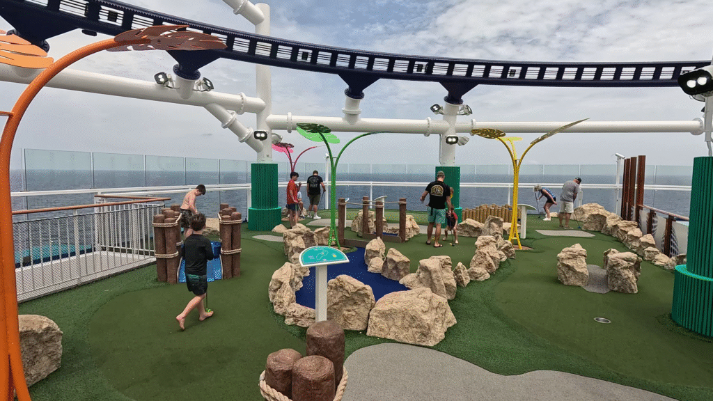 A family playing mini golf on the outdoor deck of the Carnival Jubilee cruise ship with the BOLT roller coaster track and the ocean in the background.