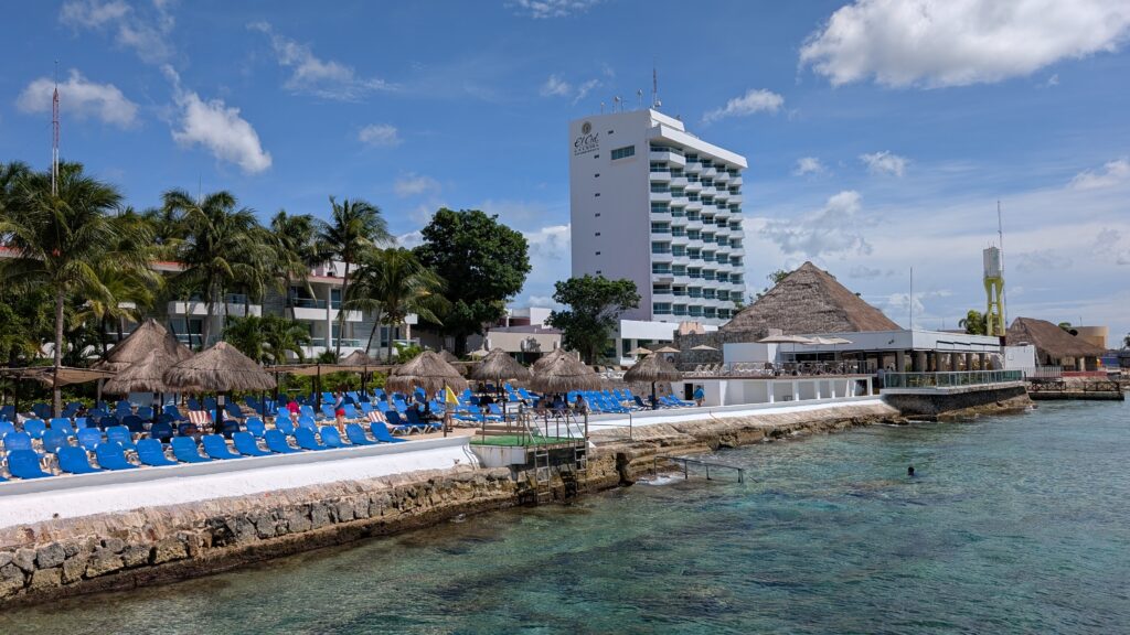 A sunny, wide-angle view of El Cid La Ceiba Beach Hotel in Cozumel, Mexico, showing blue lounge chairs lined up along a stone sea wall next to clear turquoise water, with a tall white hotel building and thatched-roof palapas under a bright blue sky with fluffy white clouds.