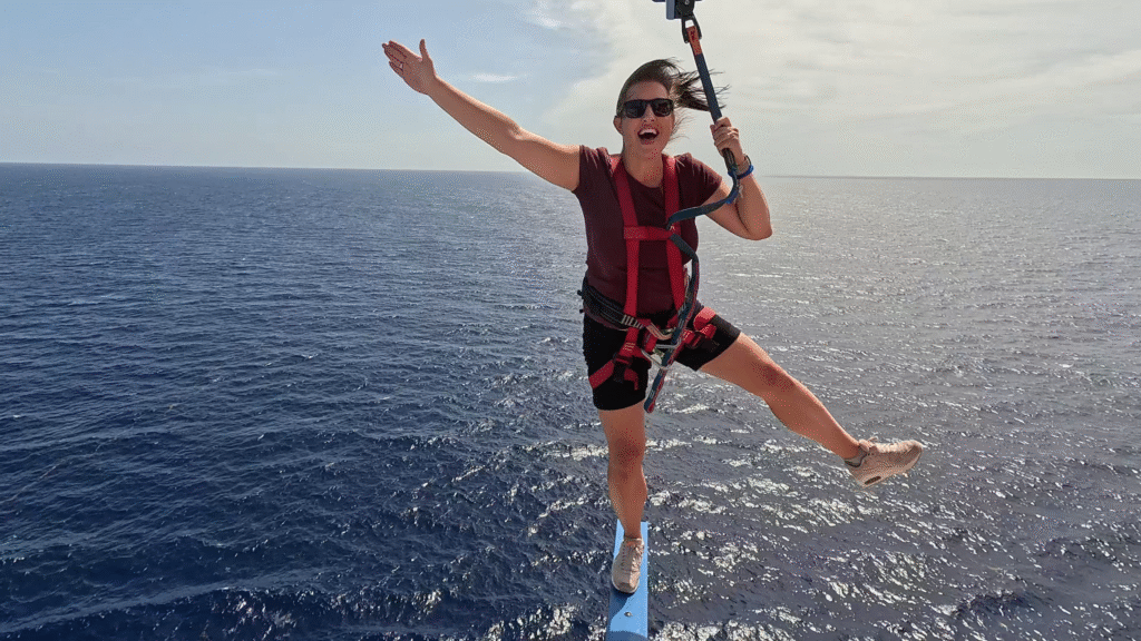 A woman wearing a red safety harness and sunglasses balances excitedly on a narrow blue beam extending out over the open ocean from the Carnival Jubilee. She has one arm raised and one leg lifted in a playful pose against a clear, sunny sky.