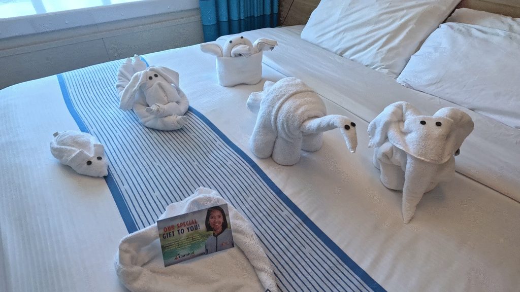 A collection of five creative towel animals, including a mouse, elephant, dinosaur, and rabbit, with black button eyes, are arranged on a white cruise ship bed with a blue and white striped runner. In the foreground is a small postcard that says "OUR SPECIAL GIFT TO YOU!" next to the folding, confirming the gesture of the cruise staff.