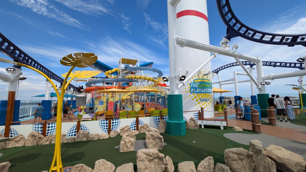 A wide-angle, sunlit view of the Deck 18 Ultimate Playground on the Carnival Jubilee cruise ship, featuring a green mini-golf course with large rocks in the foreground, the BOLT rollercoaster track and blue waterslide in the background, and white funnel with "The Ultimate Playground" sign.
