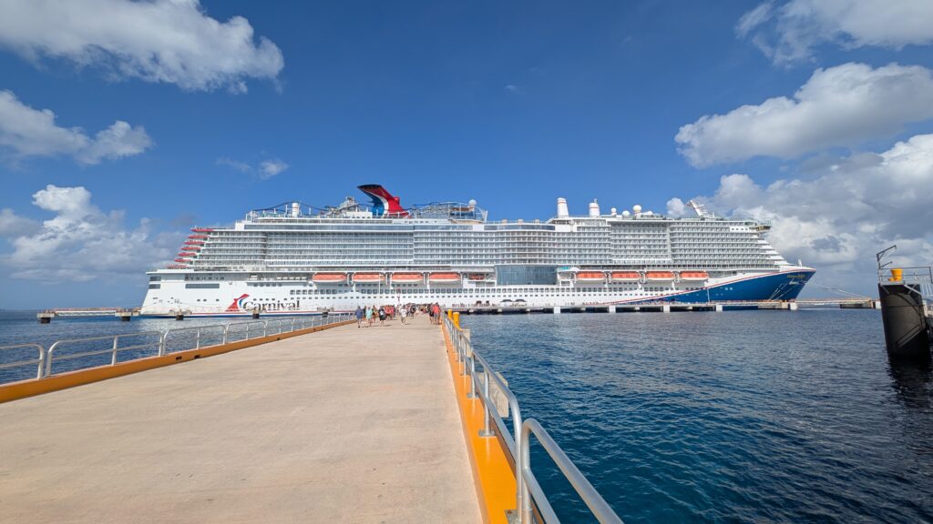 The large, multi-deck Carnival Jubilee cruise ship is docked in Cozumel, Mexico, alongside a long concrete pier under a blue sky with scattered clouds.