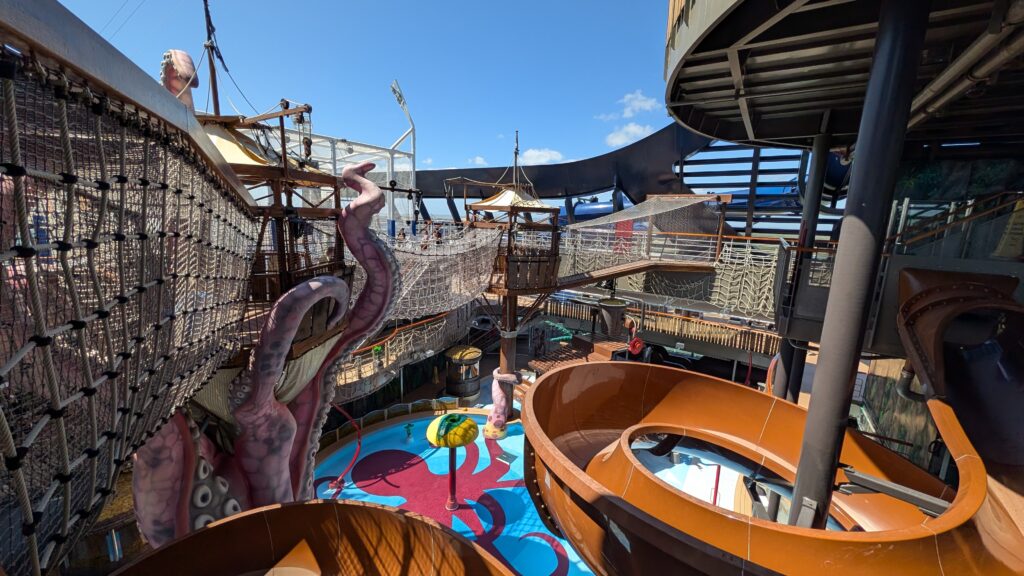 A high-angle view of Pirate’s Cove on the MSC Seascape, featuring large brown waterslides, a colorful splash pad with a yellow mushroom fountain, giant decorative pink octopus tentacles, and elevated rope bridges under a clear blue sky.