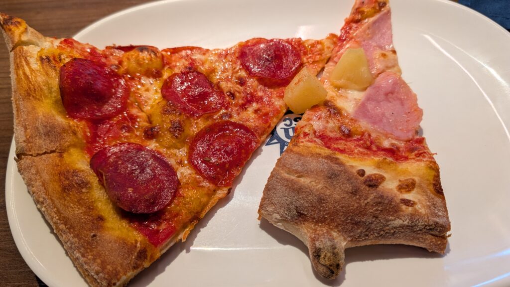 Two slices of thin-crust pizza on a white plate with the MSC logo, featuring pepperoni and ham with pineapple, from the buffet of the MSC Seascape cruise ship.