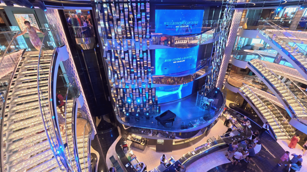 An elevated view looking down into the multi-story atrium of the MSC Seascape cruise ship. The modern, blue-lit central core features large LED screens displaying "Willkommen An Bord" (Welcome on Board), surrounded by hanging silver decorative panels. On either side are dramatic, sparkling spiral staircases lit with blue lights. At the bottom, a bar area is busy with passengers and staff, and a grand piano sits on a lower level. A passenger is walking down the left staircase.