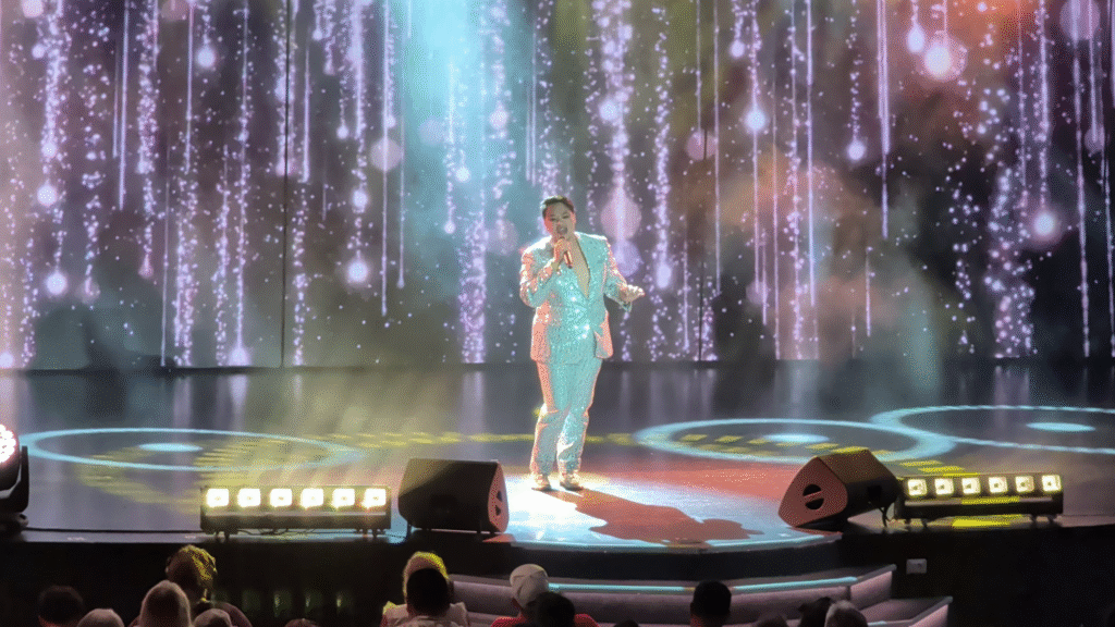 Performer singing during the MSC Seascape Crew Talent Show in the Chora Theatre, illuminated by colorful stage lighting and visual effects.