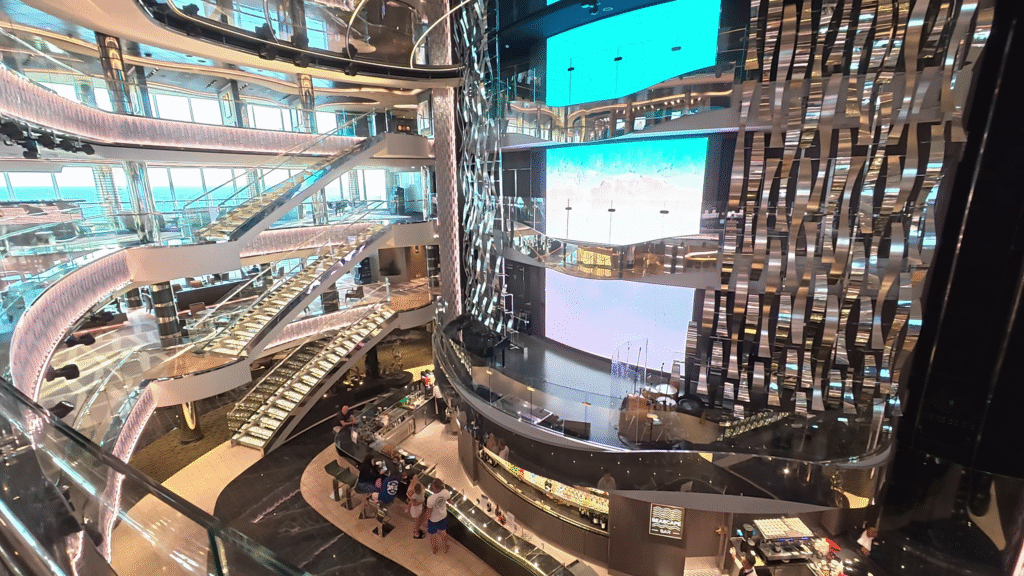MSC Seascape multi-level atrium with glass railings, large screens up the middle, a bar on the lower level, and a raised entertainment stage above in an open elegant space.