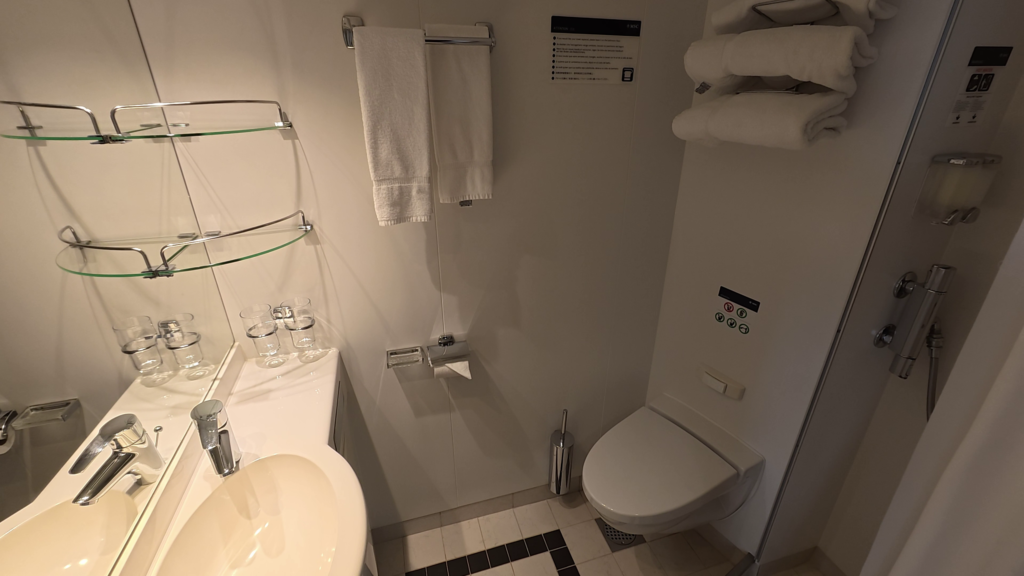Bathroom in MSC Poesia aft balcony cabin 11250 with sink, mirror, storage shelves, toilet, towels, and shower.