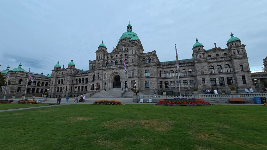 British Columbia Parliament Buildings in Victoria, BC, featuring historic stone architecture, green copper domes, lawns, and colourful flower gardens.